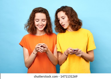 Two Identical Twins Holding Mobile Phones, Making A Phone Call, Playing Online Games, Surfing, Browsing The Net . Isolated Blue Background, Studio Shot