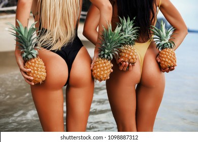 two incredibly beautiful sexy model girls in bikini at the seaside of the tropical island, blonde brunette, bronze tan, travel summer vacation, fashion style, in the hands pineapples behind the back