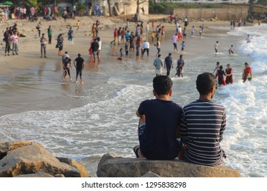 Two Indian Friends Sitting On The Rocks And Looking At People Playing On The Beach. Fort Kochi Beach.