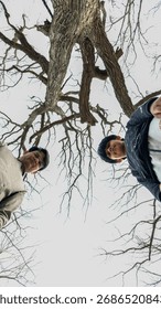 Two individuals captured from a low-angle perspective stand against the backdrop of a large, leafless tree. The intricate network of bare branches extends upwards, creating a striking pattern against 