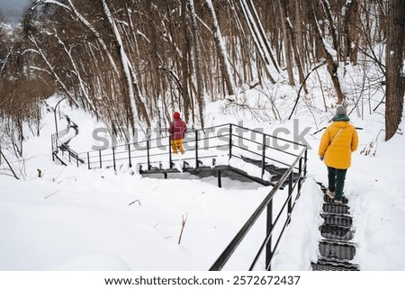 Two individuals are carefully navigating their way down a set of stairs that are covered in a blanket of snow during a chilly winter day. The environment around them feels quite frosty