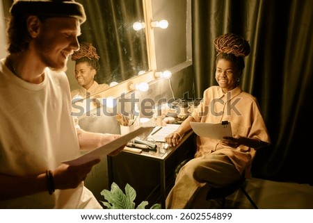 Two individuals collaborating on script in a cozy dressing room with a lighted mirror. One person seated, holding papers, while other stands and smiles