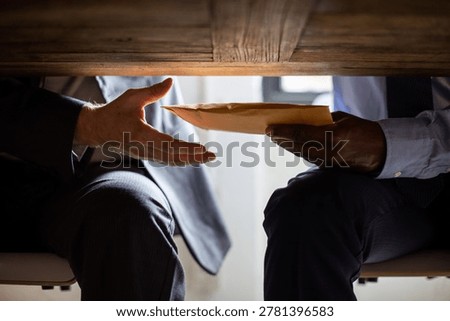 Two individuals exchange an envelope under a table, suggesting a secretive or confidential transaction in a business setting.
