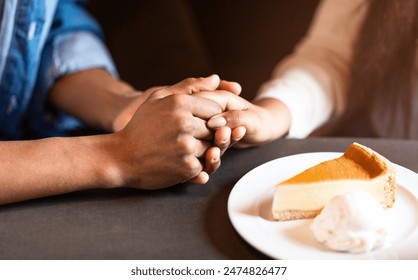 Two individuals are pictured holding hands at a table in a cozy cafe. A slice of cheesecake on a plate with a dollop of whipped cream is in front of them, suggesting a warm and intimate moment - Powered by Shutterstock