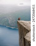 Two individuals stand on the edge of Preikestolen, a dramatic cliff in Norway, gazing out at the vast expanse of the fjord below. coupe of men and women at Preikestolen, Norway