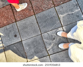 Two individuals stand on patterned pavement, showcasing white shoes against a sunlit outdoor backdrop.