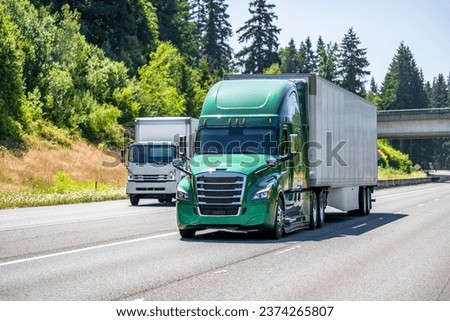 Two industrial carriers green big rig semi truck with dry van semi trailer and small rig cab over semi truck with box trailer running side by side on the multiline highway road