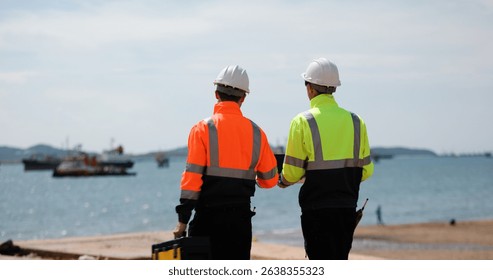 Two industrial workers wearing high-visibility safety jackets and white hard hats carry tool box maintenance operation at a  industrial port