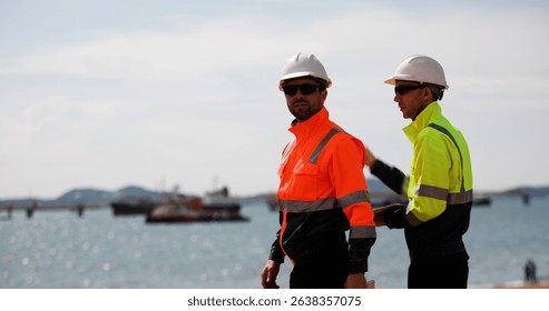 Two industrial workers wearing high-visibility safety jackets and white hard hats carry tool box maintenance operation at a  industrial port