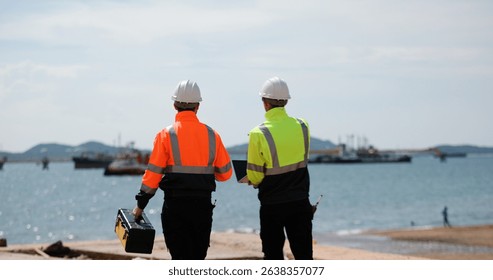 Two industrial workers wearing high-visibility safety jackets and white hard hats carry tool box maintenance operation at a  industrial port