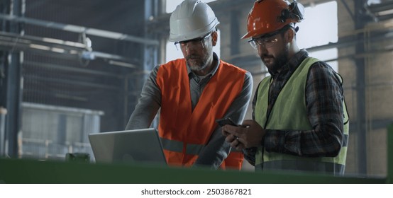 Two industry technicians in safety uniform and hard hats stand in warehouse, work on laptop computer and mobile phone. Mature engineer and adult worker talk at manufacturing factory. Slow motion. - Powered by Shutterstock