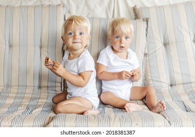 Two Innocent Babies Are Enjoying Cookies. Portrait Of Twins With Blue Eyes And Blonde Hair Eating Biscuits On A Sofa In The Living Room. Funny Picture Of A Baby, Identical Kids Eating Sweets Together