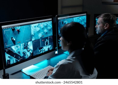 Two intercultural security guards in uniform sitting by workplace in front of computer monitors and observing cctv video on screens - Powered by Shutterstock