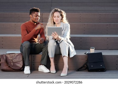 Two Intercultural White Collar Workers Looking Through Online Data On Tablet Screen At Meeting