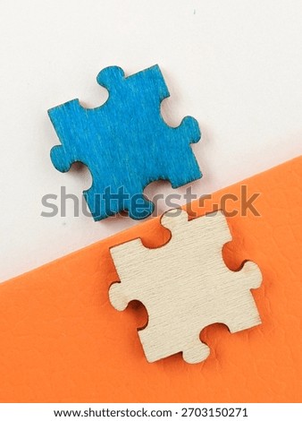 Two interlocking puzzle pieces, one blue and one natural wood, sit on a diagonal white and orange surface. The image evokes connection, teamwork, problem solving and creative collaboration