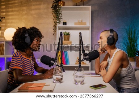 Two interracial content creators are sitting in a home recording studio with headphones on and talking at the microphone while discussing topics. Two girls are live streaming an episode for internet.