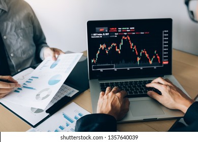 Two Investors Are Working Together With Analyzing The Stock Data Graphs In The Paper And Viewing The Data On The Laptop Screen.