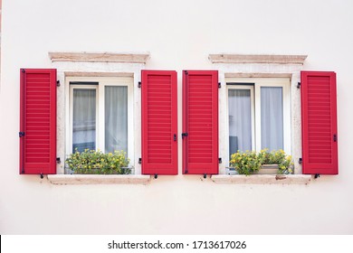 Two Italian windows on the white wall facade with open red color classic shutters and flowers on the windowsill