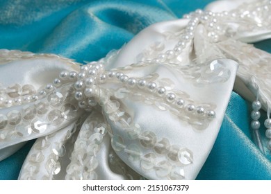 Two ivory white satin beaded bows for wedding dress trim decorated with faux pearls and sequins, arranged on turquoise satin fabric.