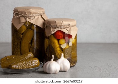 Two jars of pickled cucumbers and assorted vegetables. Fresh garlic and pickles on a gray table. Copy space