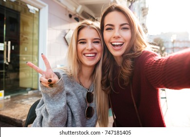 Two joyful attractive girls taking a selfie while sitting together at cafe and showing peace gesture outdoors