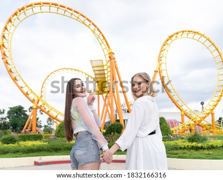 Two joyful cheerful girls, holding hands together, smiling and looking to camera at the amusement theme park on Roller coaster background. tourism, people, holiday concept