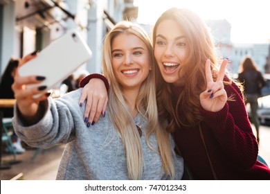 Two joyful cheerful girls taking a selfie while sitting together at cafe and showing peace gesture outdoors