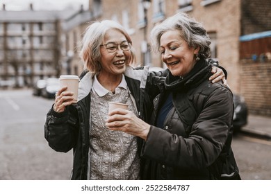 Two joyful diverse older women friends share a warm moment outdoors, laughing and enjoying coffee together. Their friendship shines as they bond over coffee. Haliday with friend concept - Powered by Shutterstock