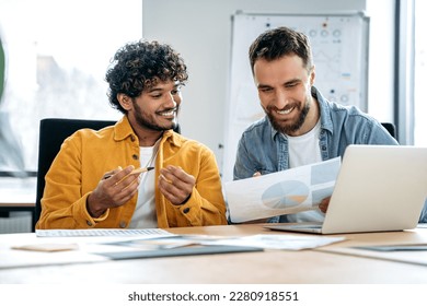 Two joyful male multiracial employees sit in the office at the table, look at the laptop screen and documents, work on a joint project, consult each other, discuss ideas and strategies, evaluate risks - Powered by Shutterstock