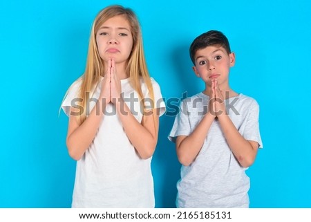 two kids boy and girl standing over blue background keeps palms pressed together in front of her having regretful look, asking for forgiveness. Forgive me please.