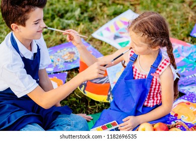 Two Kids Brither And Sister In Blue Aprons Tickle Nose Each Other By Paint Brushes, Sitting Outdoor In Autumn Time On The Grass. Learning Painting Drawing Art School Concept.