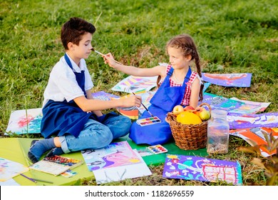 Two Kids Brither And Sister In Blue Aprons Tickle Nose Each Other By Paint Brushes, Sitting Outdoor In Autumn Time On The Grass. Learning Painting Drawing Art School Concept.