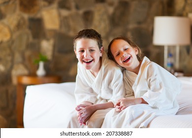 Two Kids Brother And Sister Wearing Little Bathrobes Laughing While Sitting On Bed At Hotel Room Or Home