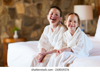 Two Kids Brother And Sister Wearing Little Bathrobes Laughing While Sitting On Bed At Hotel Room Or Home