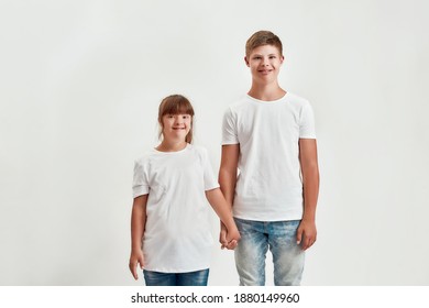 Two kids, disabled boy and girl with Down syndrome smiling at camera, holding each other hands while posing together isolated over white background. Children with disabilities and special needs