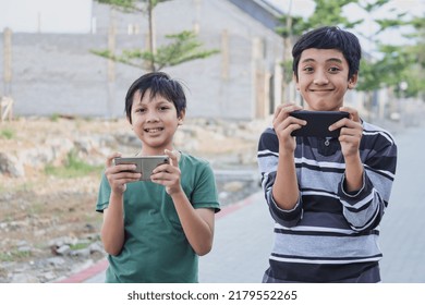 Two Kids With Gadgets. Brothers Surfing The Net Or Playing Online Games On Smartphone And Digital Tablet Outside While Standing. Modern Communication And Gadget Addiction Concept.