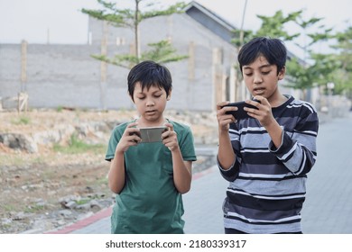 Two Kids With Gadgets. Brothers Surfing The Net Or Playing Online Games On Smartphone And Digital Tablet Outside While Standing. Modern Communication And Gadget Addiction Concept.
