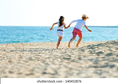 Two Kids Holding Hands Running Away On Sandy Beach.