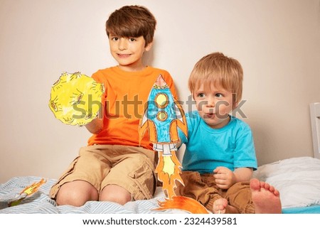 Two kids play astronauts holding paper rockets and planets at home with little boy show expression for liftoff