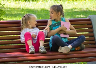 Two Kids Quarrel On A Bench In The Park. The Older Sister Does Not Want To Share Books With A Younger And Puts Out One's Tongue