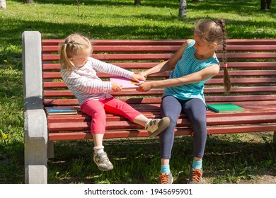 Two Kids Quarrel On A Bench In The Park. Two Little Girls Do Not Want To Share And Are Fighting Over A Book