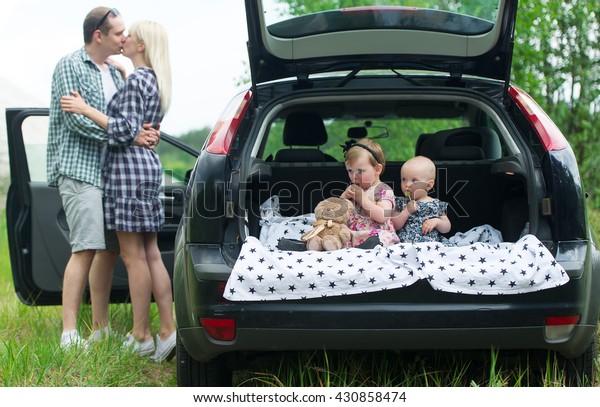 Two kids sit in a car luggage\
carrier. Near the car there are parents of kids. They\
kiss.