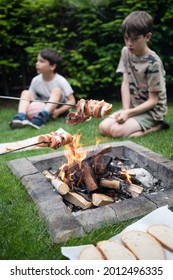 Two Kids Slices Of Sausage, Bacon And Onion  Roasted On Bonfire Flame With Blurred Kids In Background. Picnic, Barbecue, Cooking Food Concept. Summer Camping, Hiking, Vacation