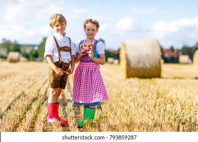 Two kids in traditional Bavarian costumes in wheat field. German children eating bread and pretzel during Oktoberfest. Boy and girl play at hay bales during summer harvest time in Germany