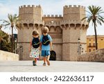 Two kids travelers walk together in hot Valencia in front of famous Torres de Serranos during their summer holidays in Spain, view from behind