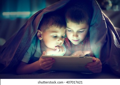 Two Kids Using Tablet Pc Under Blanket At Night. Brothers With Tablet Computer In A Dark Room