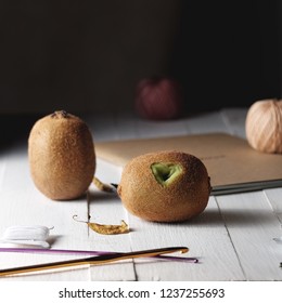 Two kiwis on a white table next to tools for knitting. On one kiwi cut 