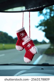 Two knitted red socks, complete with white accents, hang from a car rearview mirror, adding a festive and cozy touch to the vehicle interior aesthetic.