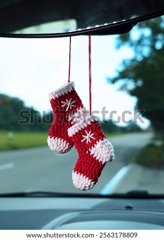 Two knitted red socks, complete with white accents, hang from a car rearview mirror, adding a festive and cozy touch to the vehicle interior aesthetic.