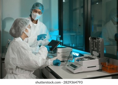 Two lab technicians in protective suits operate a moisture analyzer and tablet in a sterile lab, ideal for pharmaceutical and food quality control concepts.